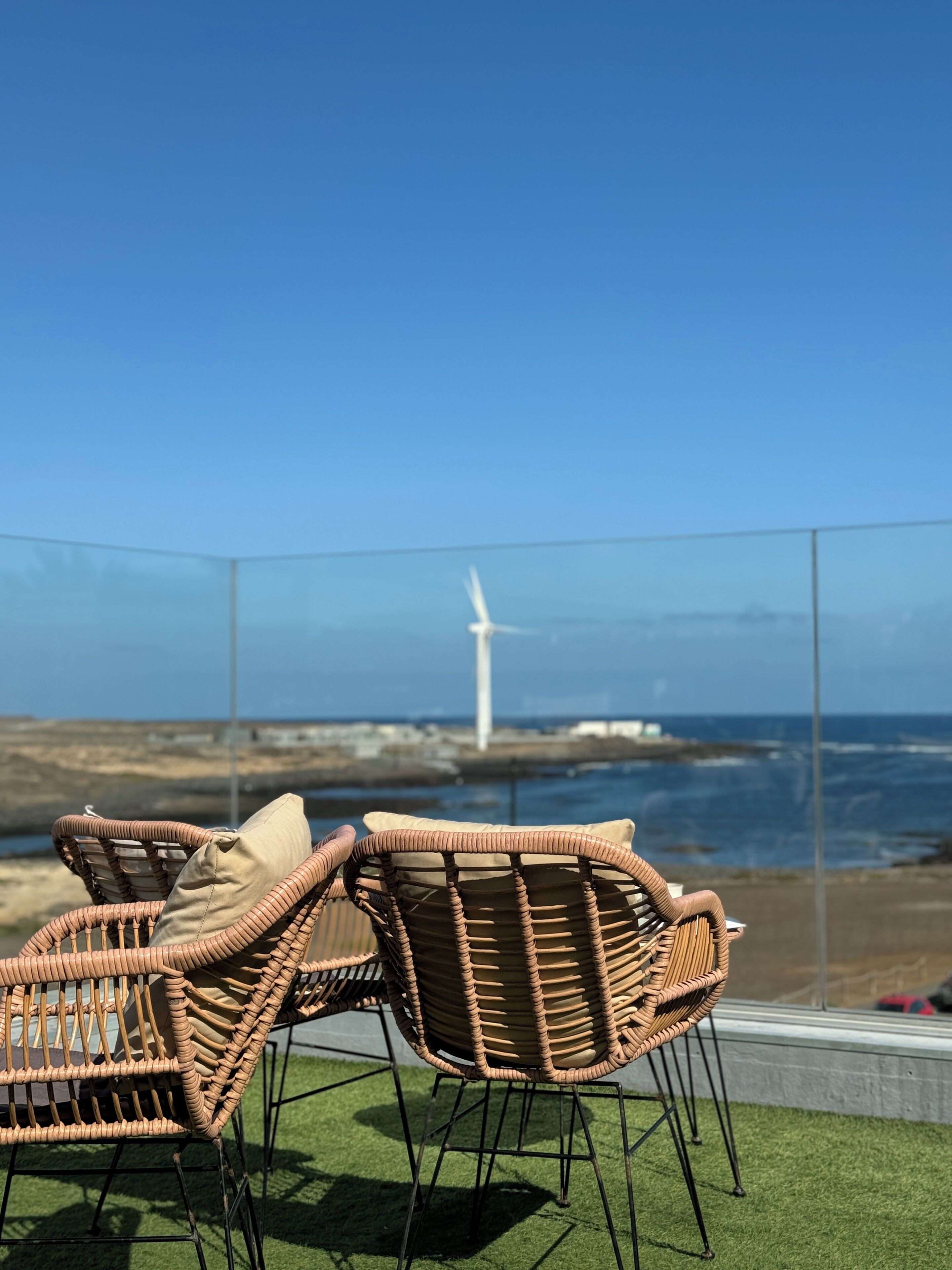 Dos sillas de mimbre con cojines en un balcón con vista a una turbina eólica y al mar.