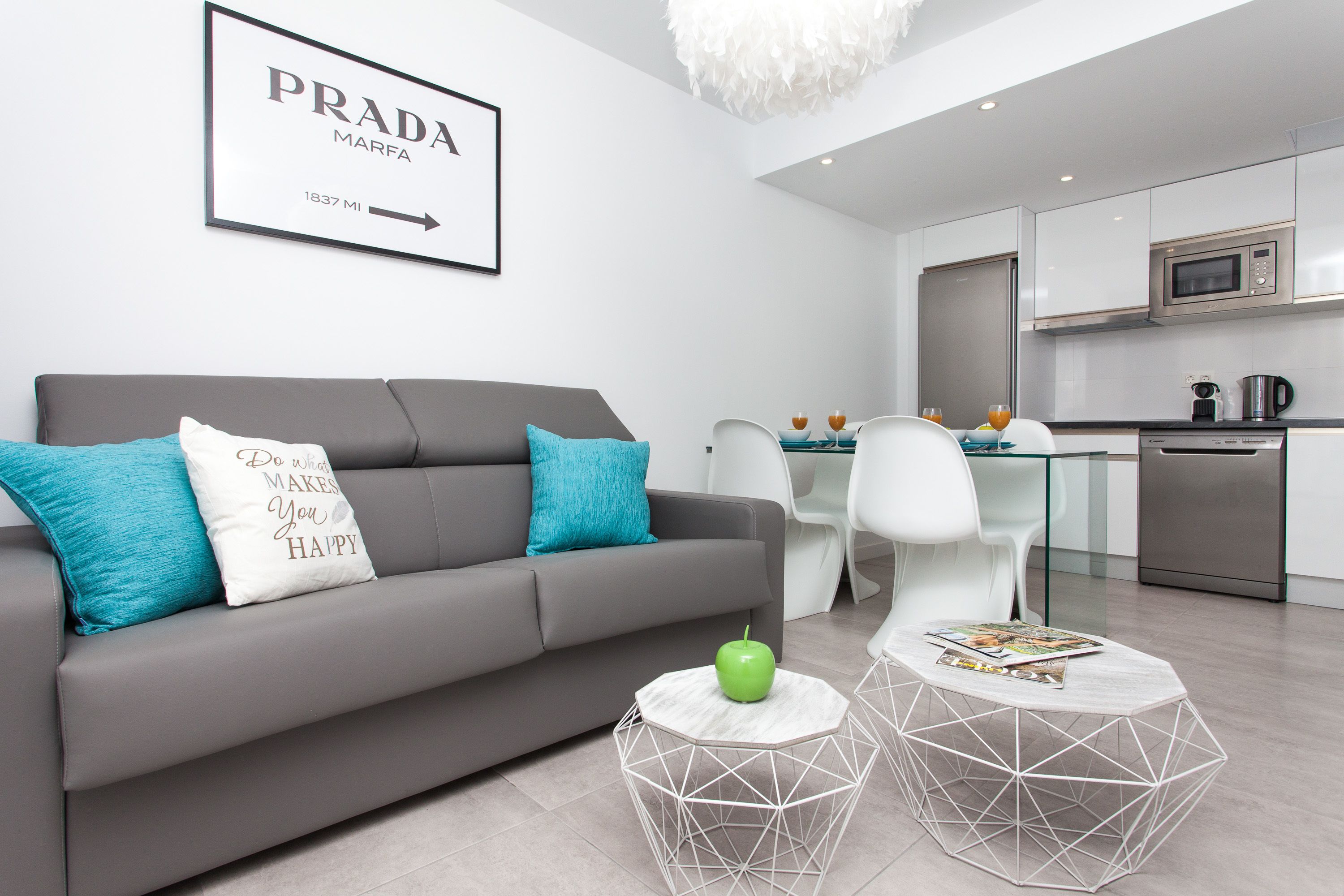 A modern living room with a grey sofa, teal pillows, and a white coffee table with a green apple. A framed sign on the wall reads "PRADA MARFA 1837 MI."