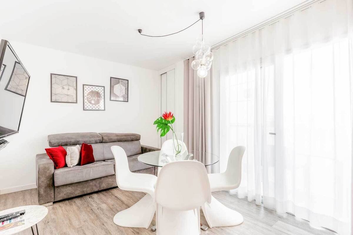 Una sala de estar moderna con un sofá gris, sillas blancas, una mesa de centro de vidrio y un televisor montado en la pared.