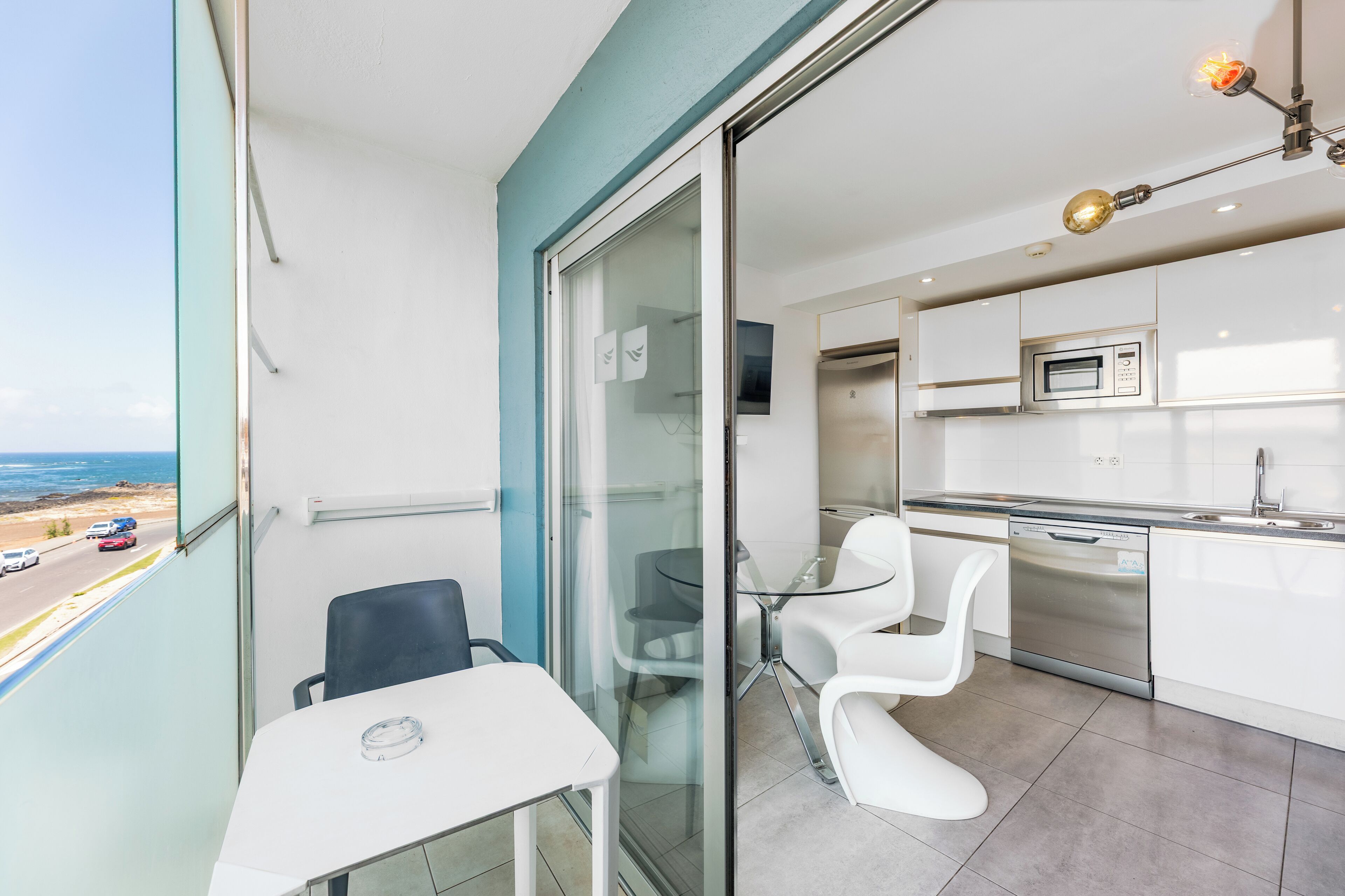 A modern kitchen with white cabinets, a stainless steel dishwasher, and a glass door leading to a balcony with a view of the sea.