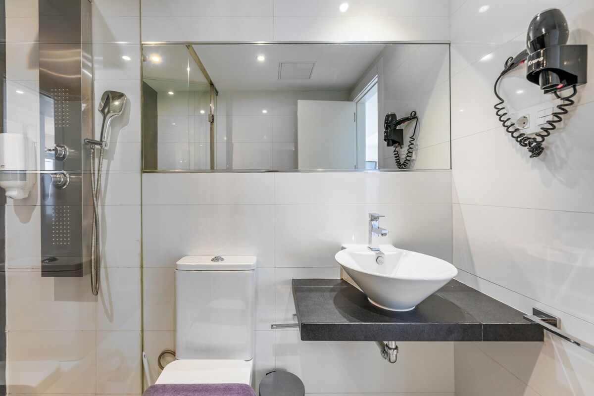 A modern bathroom with a glass-enclosed shower, a white toilet, a black countertop sink, and a wall-mounted hairdryer.