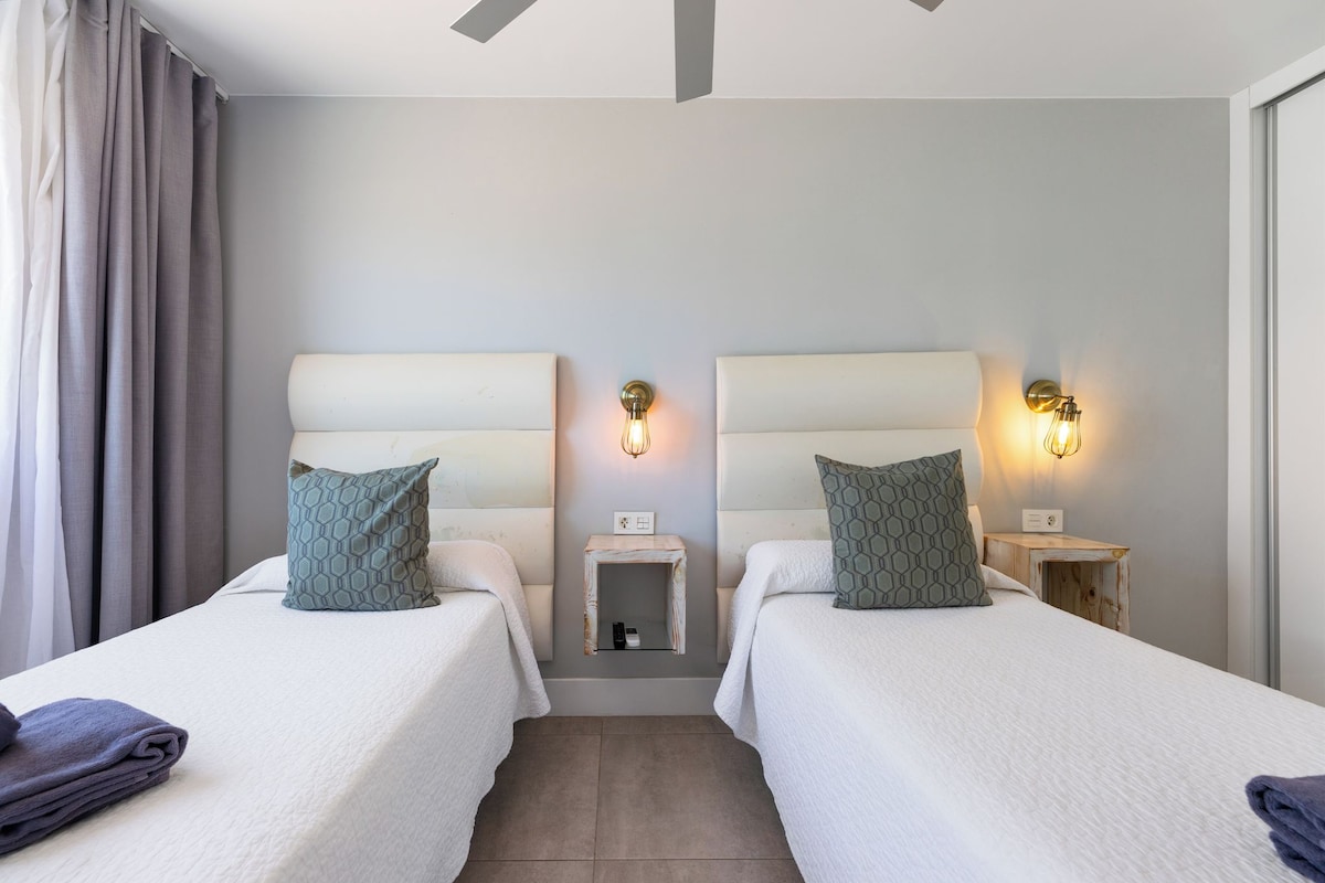 Habitación de hotel con dos camas, una mesita de noche pequeña, una lámpara de pared y un ventilador de techo.