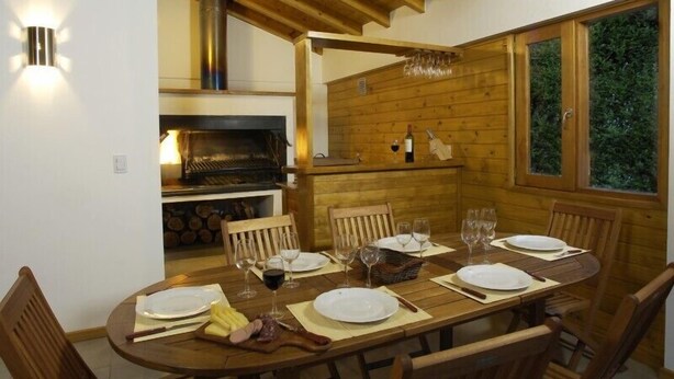 Mesa de madera para seis personas dispuesta con platos, cubiertos y vasos, un banco de madera para sentarse, una parrilla empotrada y una ventana con vistas a zonas verdes.