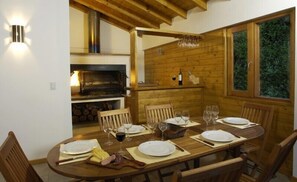 Mesa de madera para seis personas dispuesta con platos, cubiertos y vasos, un banco de madera para sentarse, una parrilla empotrada y una ventana con vistas a zonas verdes.