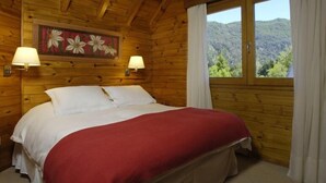 Un dormitorio de cabaña de madera con una cama, dos lámparas fijadas a la pared, un ventanal con cortinas y un cuadro floral en la pared.