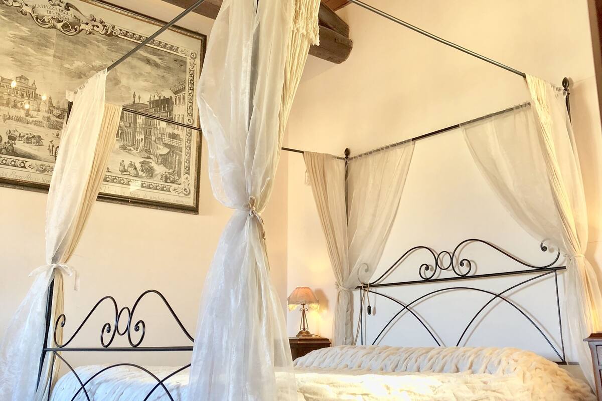 A four-poster bed with white bedding and a canopy, a chandelier, and a framed picture on the wall.