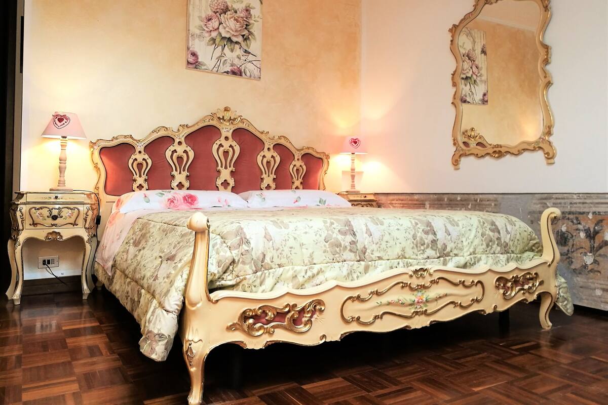 A classic four-poster bed with ornate gold detailing, flanked by nightstands with lamps, in a room with a wooden parquet floor and floral wallpaper.