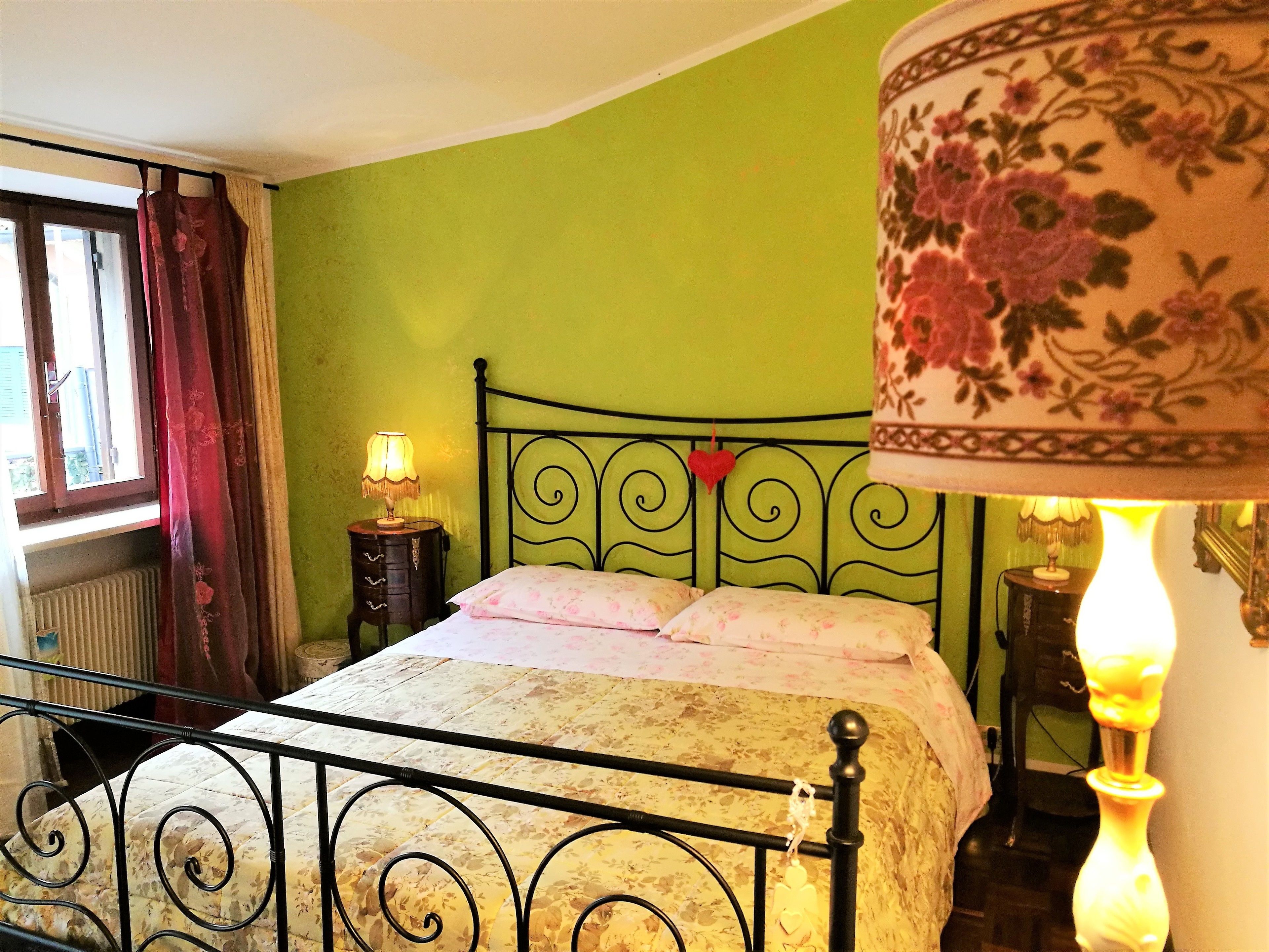 A bed with a floral bedspread, a nightstand with a lamp, a decorative heart hanging on the headboard, and a window with curtains.