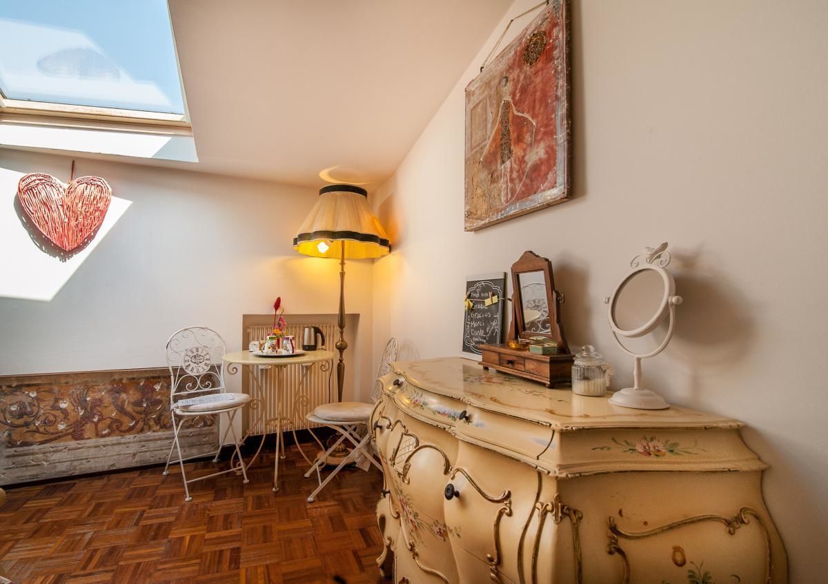 A cozy room with a wooden floor, a white chair, a table with a lamp, and a vintage dresser with a mirror.