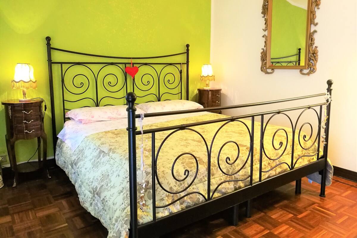 A bed with a metal frame and floral bedding, two bedside tables with lamps, a mirror, and a green wall.