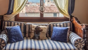 Ein gemütliches Zimmer mit Holzbalkendecke, einem fenster mit Vorhängen, einem gestreiften Sofa mit Kissen und Blick auf ein Gebäude mit einem Turm.
