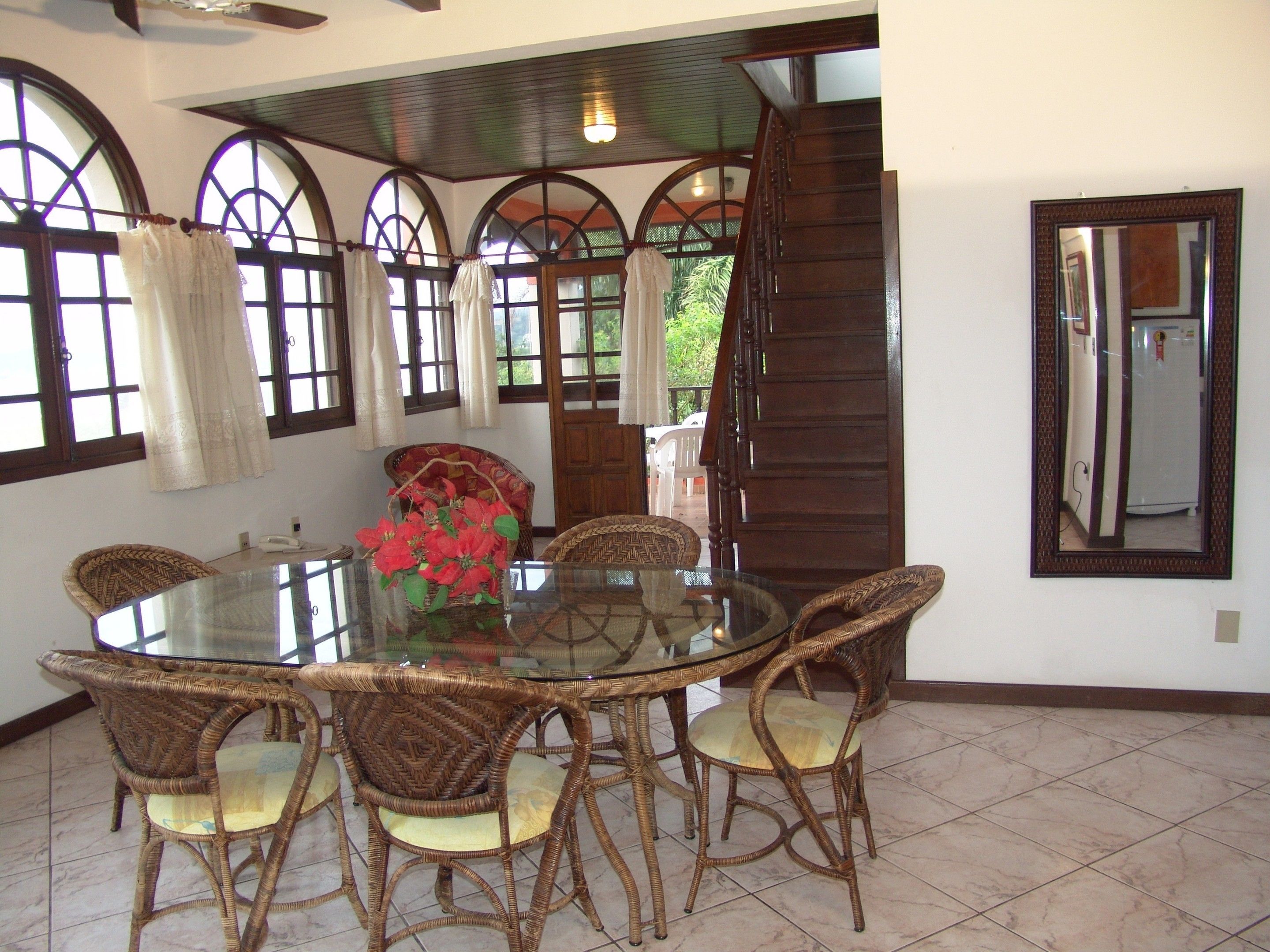 A dining area with a glass-top table surrounded by wicker chairs, a vase with red flowers, and a large mirror on the wall.