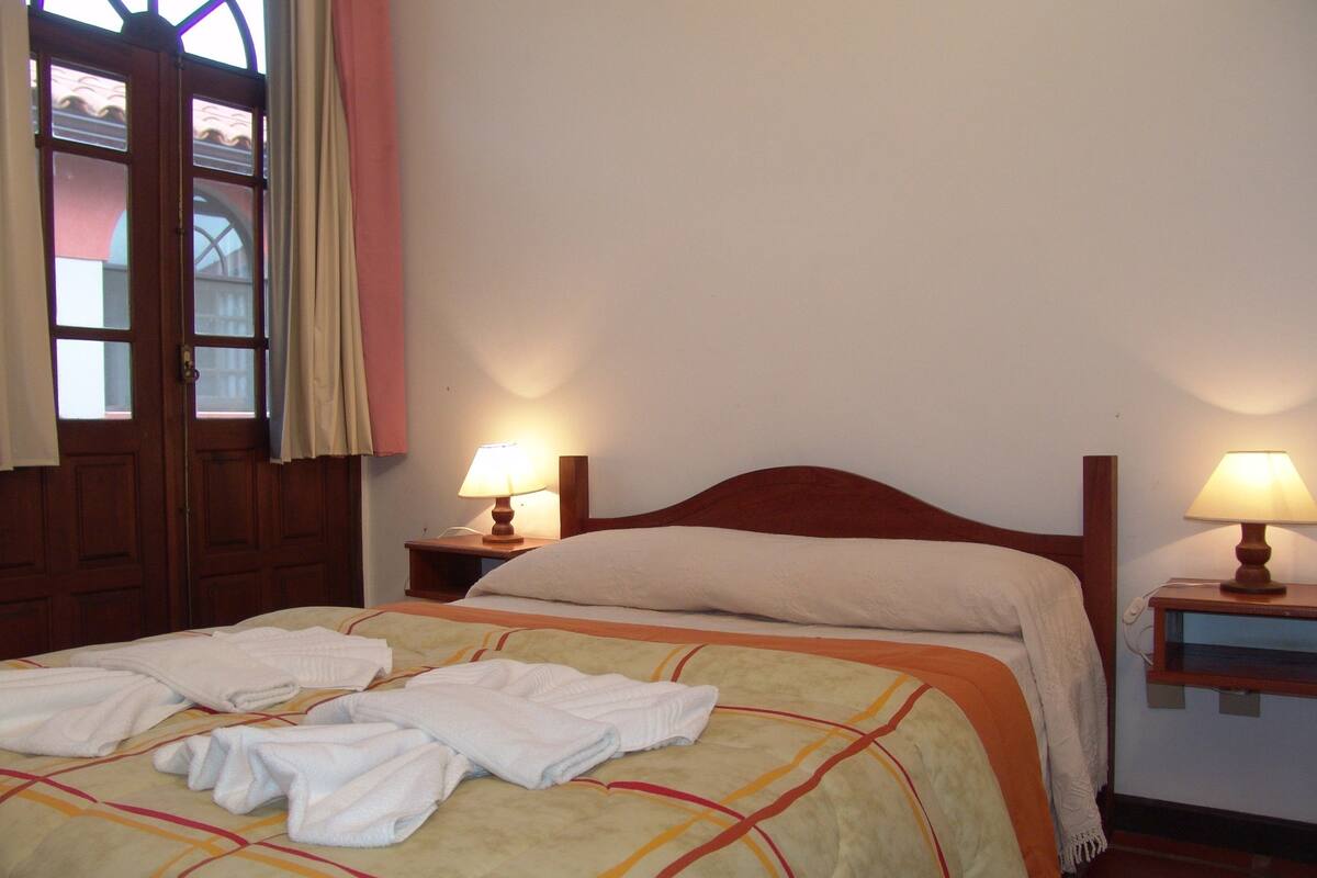 Una habitación de hotel con cama, dos lámparas de noche, una ventana con cortinas y una puerta.