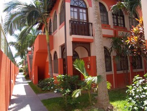 Un edificio con facciata rosso e bianco, numerosi balconi, e un vialetto fiancheggiato da palme e arbusti.