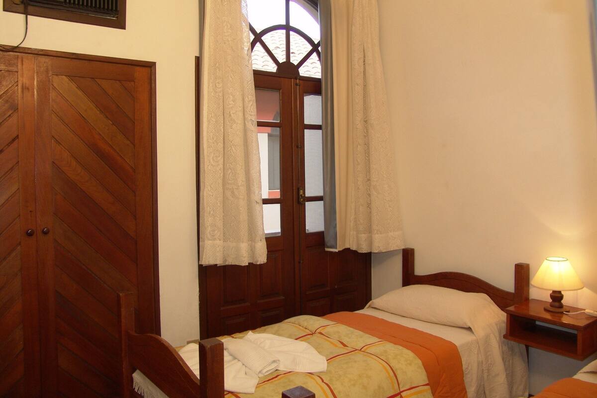 Un dormitorio con puerta de madera, ventana con cortinas, mesita de noche con lámpara y cama con manta.