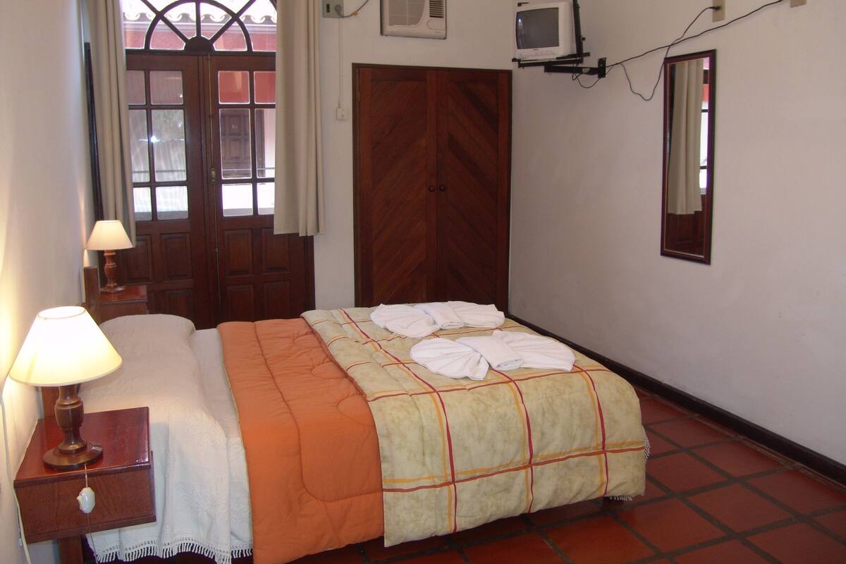Un dormitorio con cama, mesita de noche, lámpara, armario de madera, televisor y espejo.