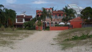 Un edificio grande de varios pisos con fachada roja y techo de tejas, rodeado de palmeras y un sendero arenoso que lleva a la entrada.