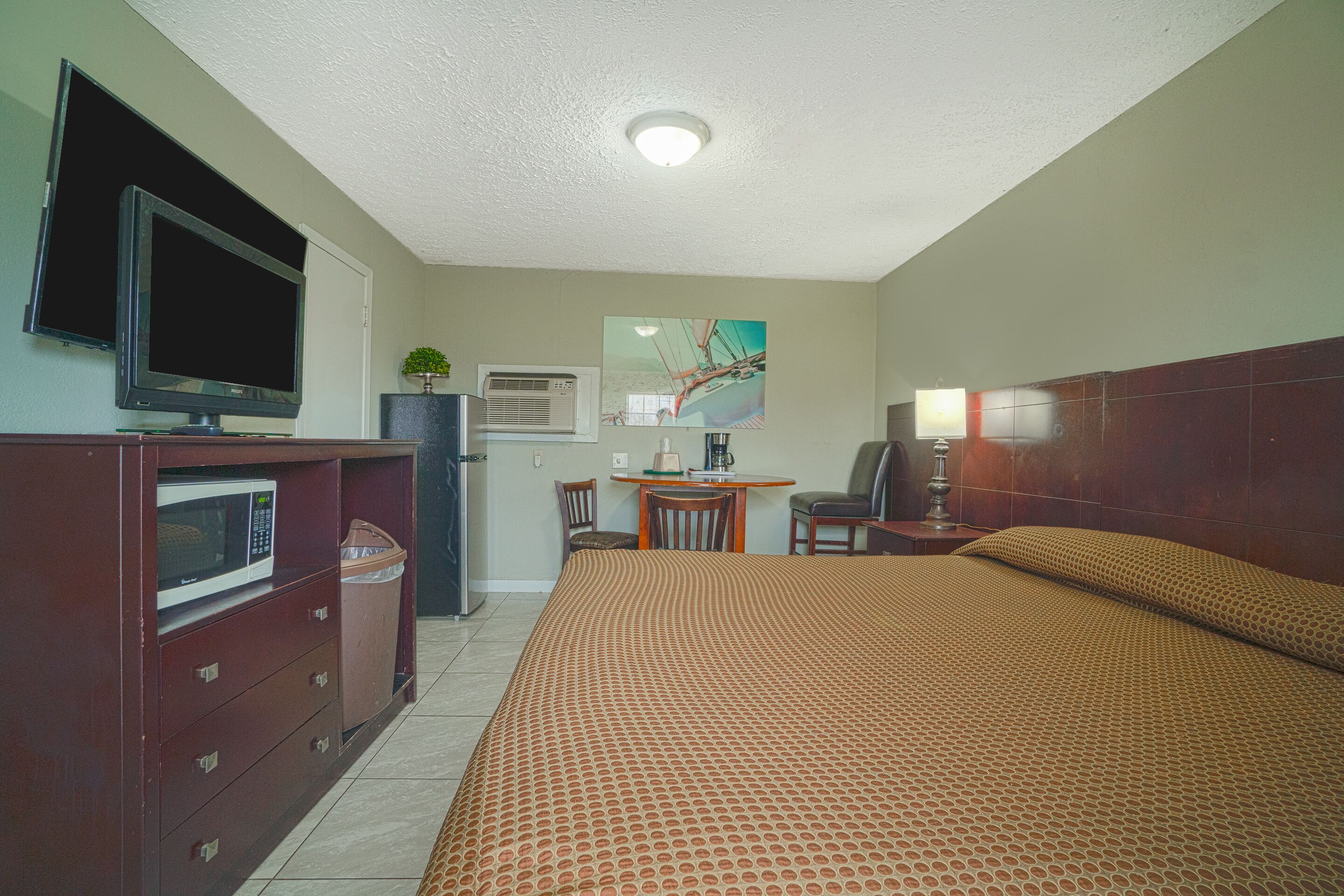 A hotel room with a bed, a TV, a desk with a chair, and a small table with a lamp.