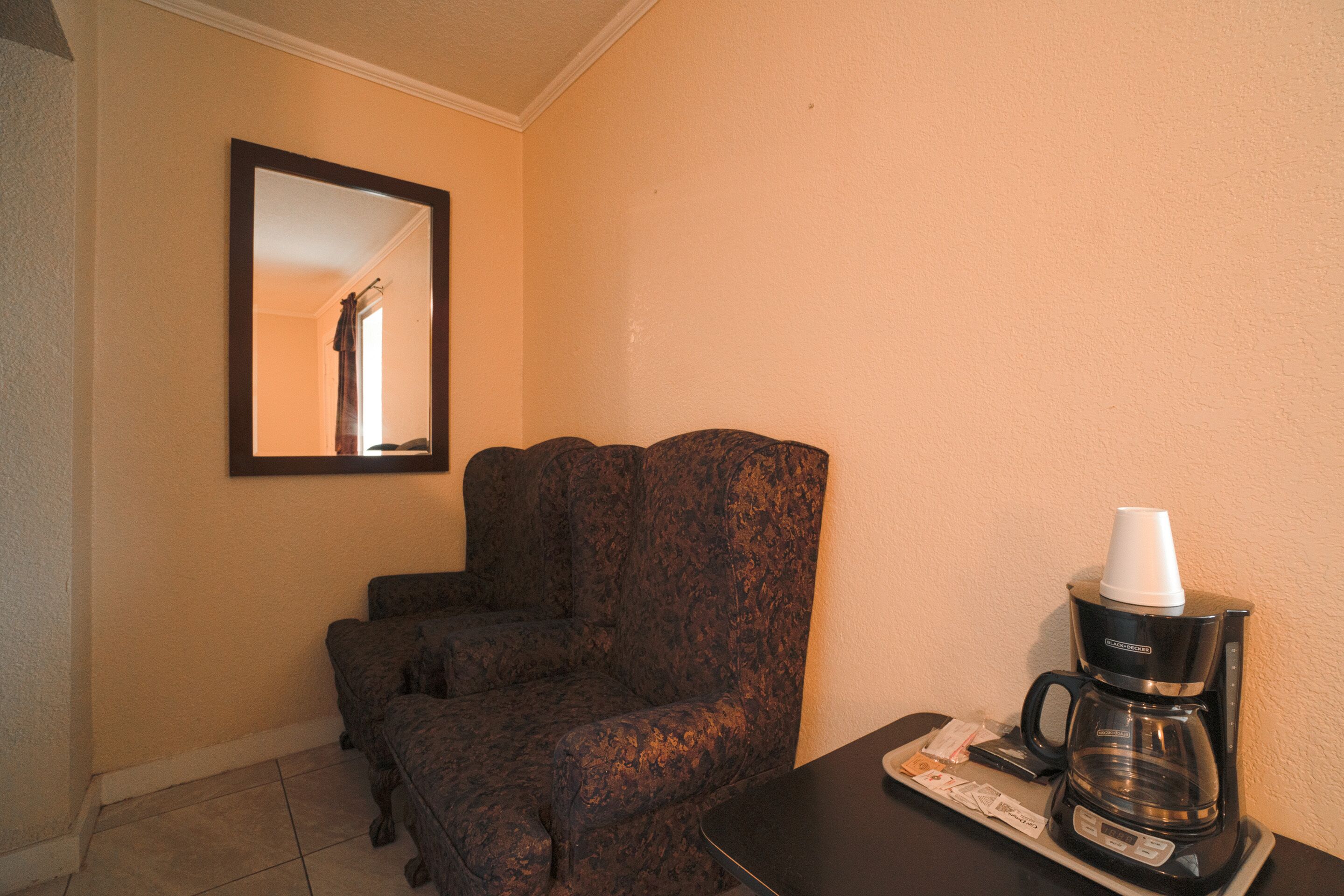 A hotel room with a dark patterned armchair, a coffee maker on a tray, and a framed mirror on the wall.