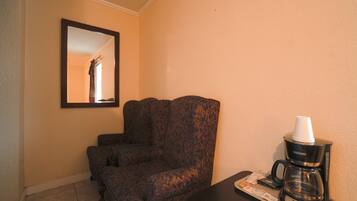 A hotel room with a dark patterned armchair, a coffee maker on a tray, and a framed mirror on the wall.