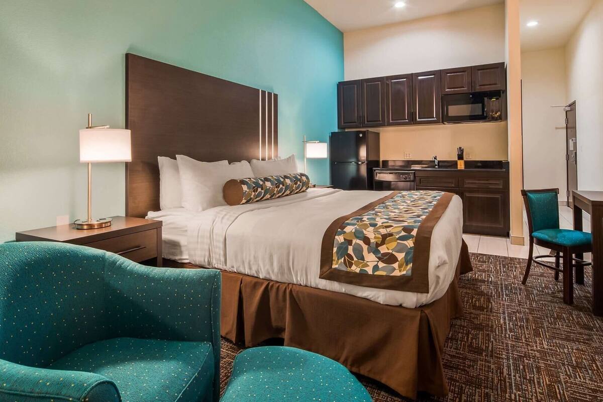 A hotel room with a bed, two chairs, a desk, a lamp, and a kitchenette.
