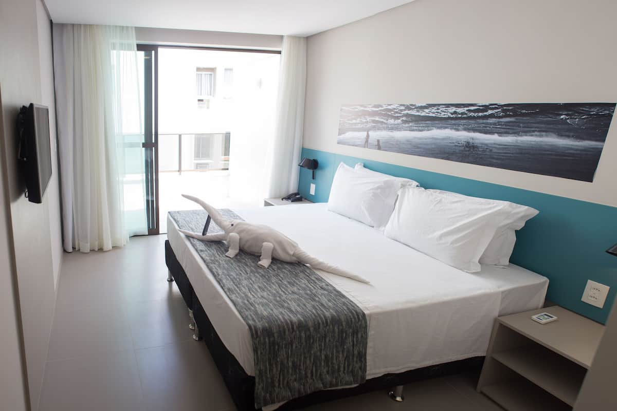 A hotel room with a bed, a nightstand, a phone, a painting of a seascape, and a view of the outside balcony.