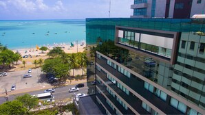 A modern building with a glass facade overlooking a beach with sailboats and parked cars. - Acqua Suites (Maceió)