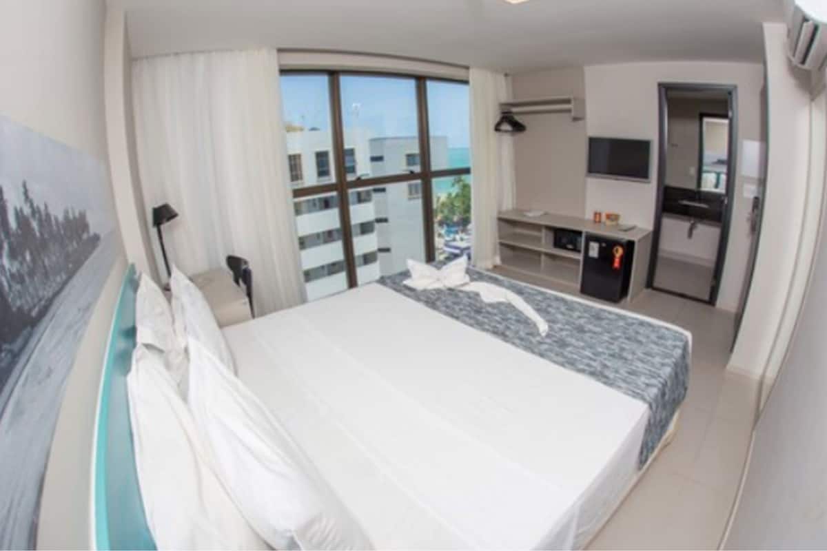 A hotel room with a large bed, a desk with a computer, a TV, and a view of the city through large windows.