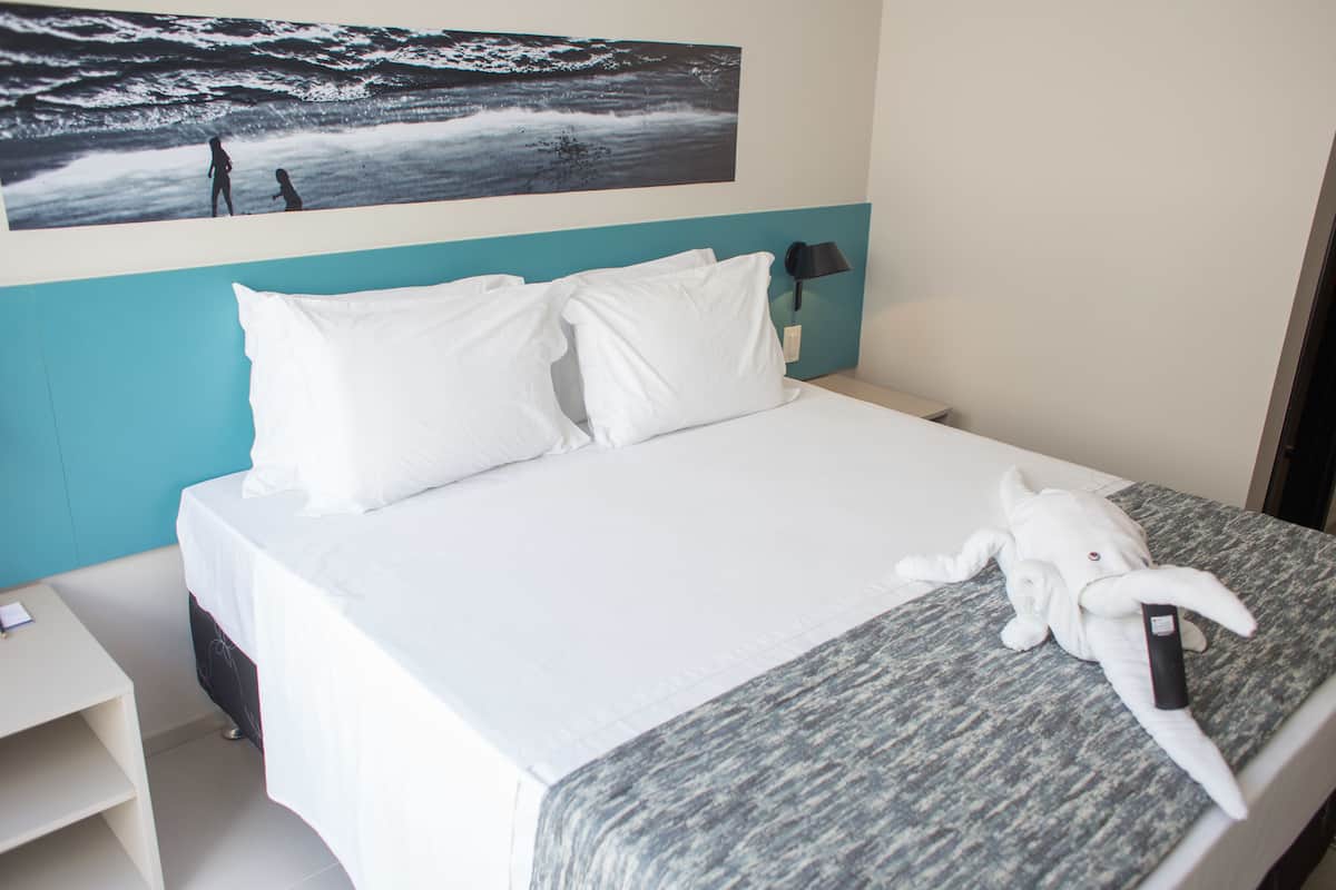 A hotel room with a bed, white pillows, a blanket, a wall-mounted painting, a nightstand, and a lamp.