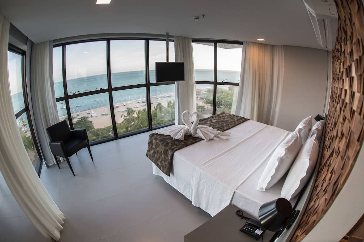 A modern hotel room with a large curved window offering a view of the beach and ocean.