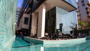 A modern building with a glass facade, a swimming pool, and outdoor seating. - Acqua Suites (Maceió)