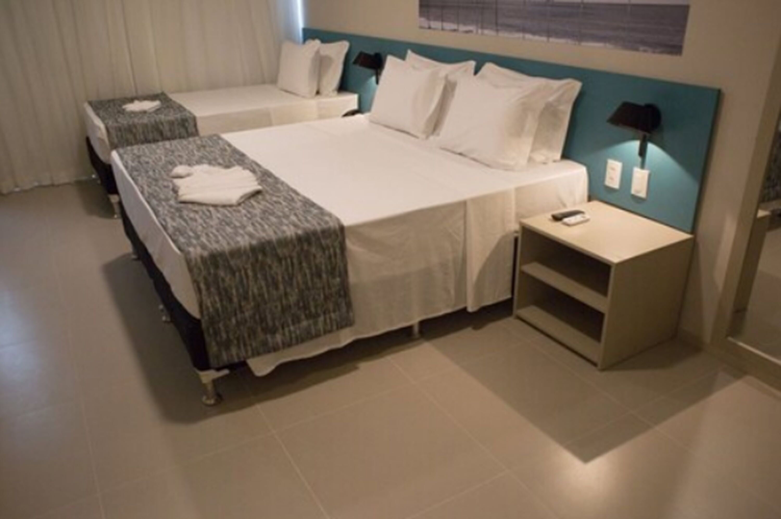 A hotel room with two single beds, a nightstand with a lamp, and a small shelf.