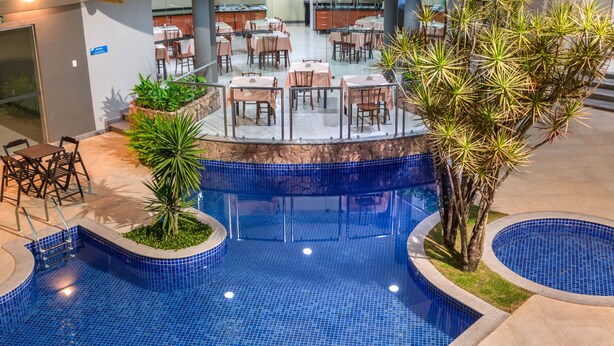 An indoor swimming pool with blue tiles, surrounded by a stone deck and palm trees. - Hotel Porto Nápolis (Eunapolis)