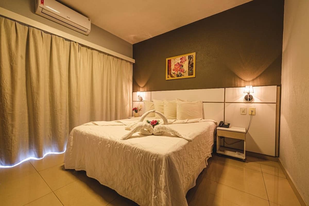 Un cuarto de hotel con una cama grande, una mesita de noche, aire acondicionado, un cuadro en la pared y una lámpara sobre la mesita.