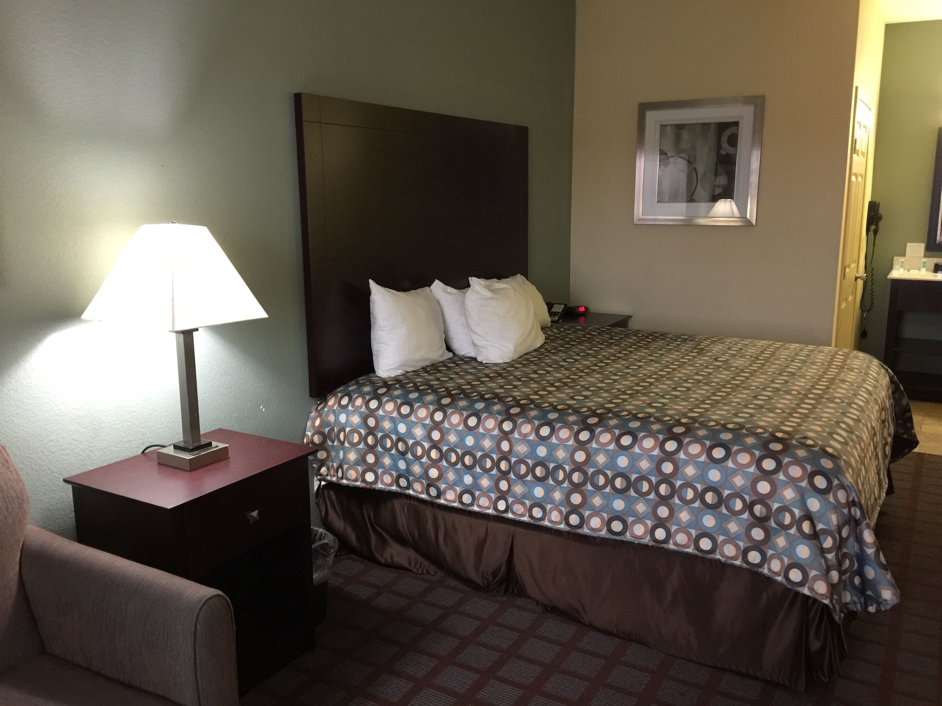 Una habitación de hotel con cama, mesita de noche, lámpara, silla y un cuadro en la pared.