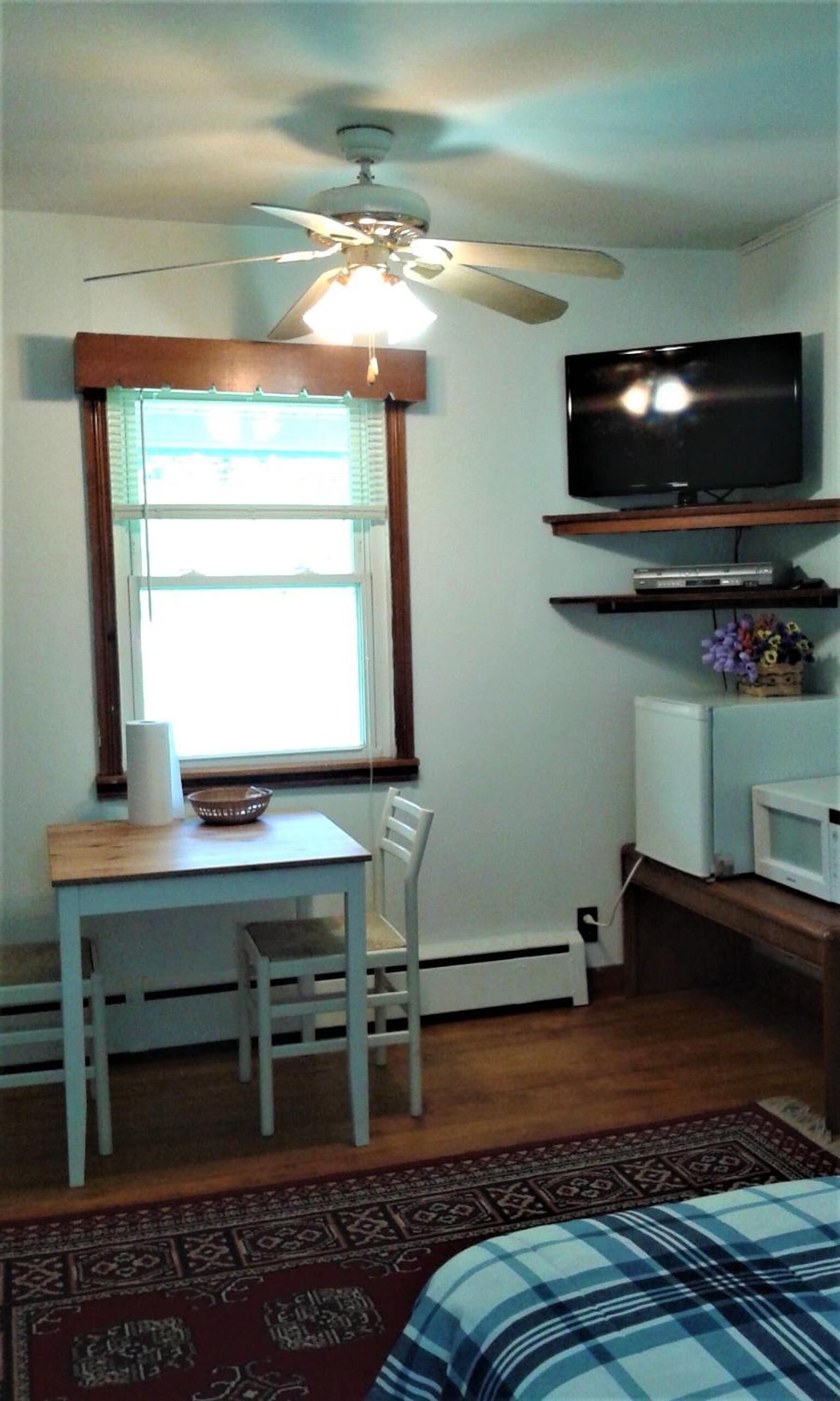 A room with a ceiling fan, a television on a shelf, a dining table with chairs, and a window with blinds.