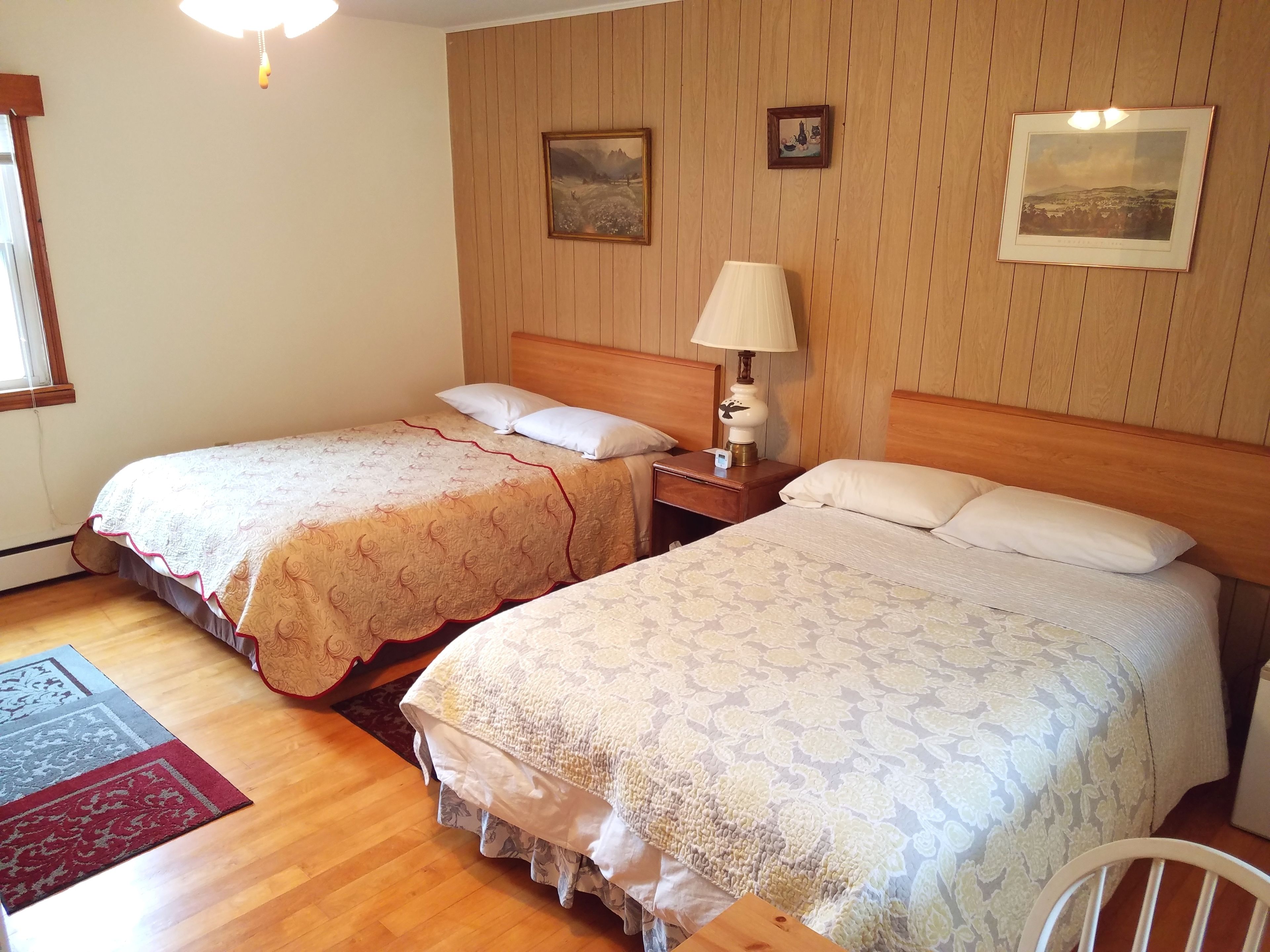 A hotel room with two beds, wooden headboards, a nightstand with a lamp, and two framed pictures on the wall.