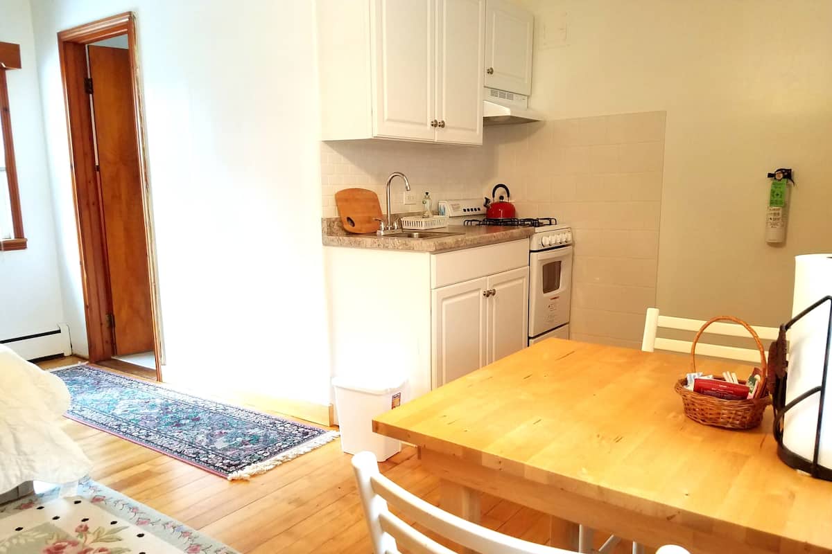 A compact kitchen with white cabinets, a wooden dining table, and a red kettle.