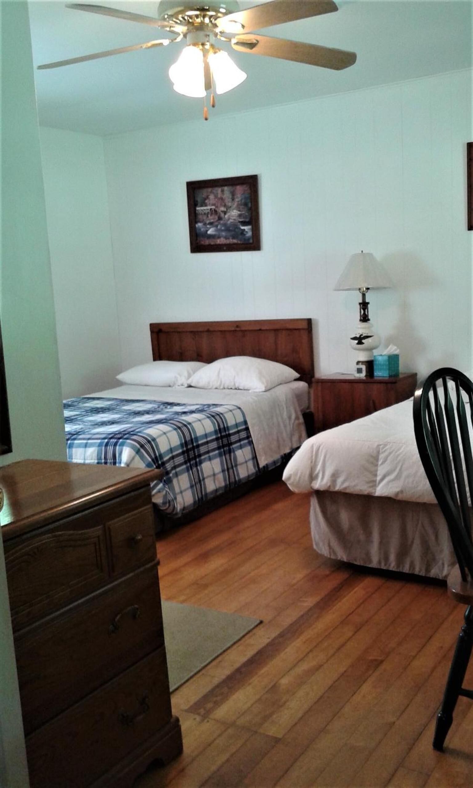 Un dormitorio con dos camas, un aparador de madera, una mesita de noche con lámpara y un ventilador de techo.