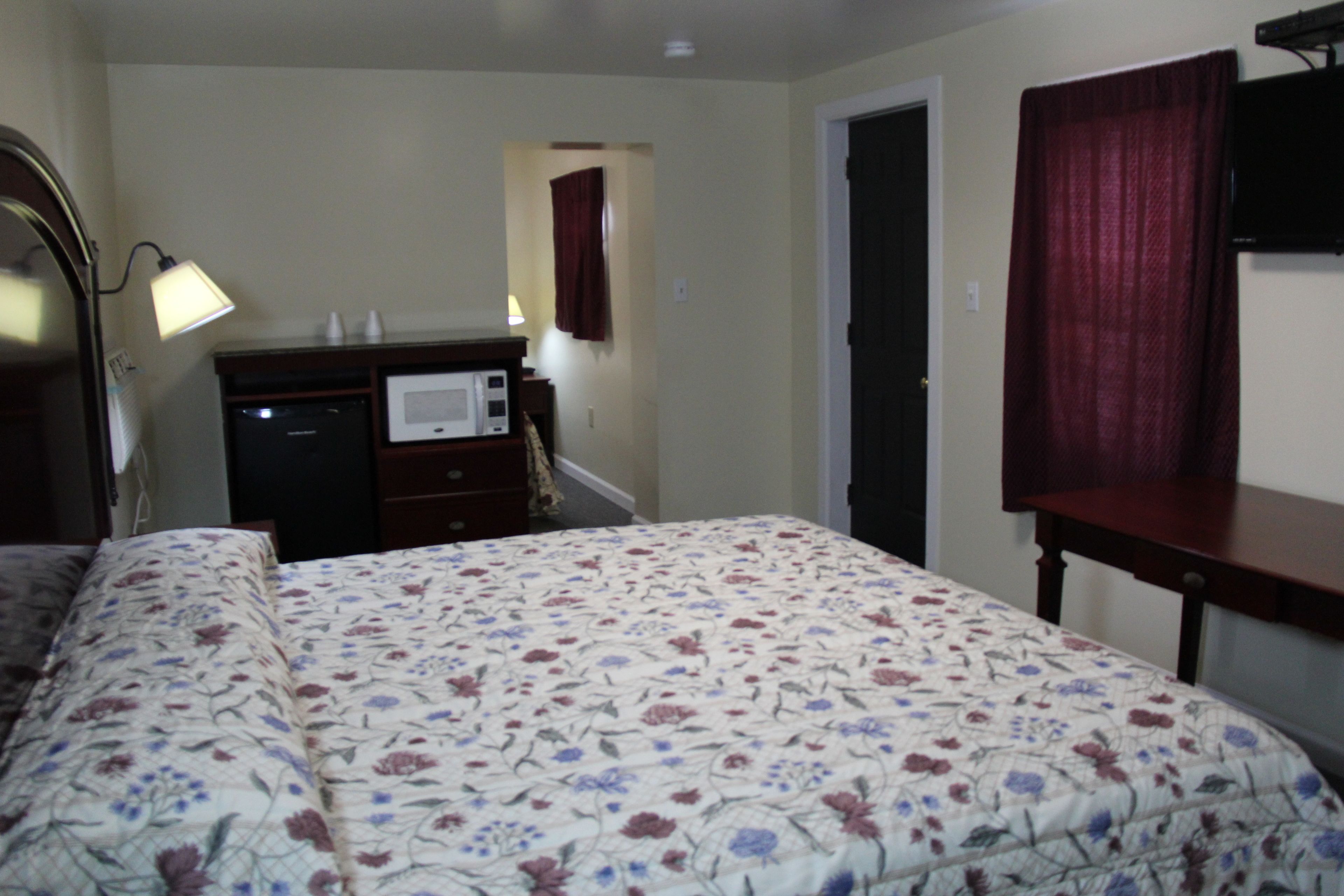 A bedroom with a floral bedspread, a bedside lamp, a dresser with a microwave, and a TV on a stand.