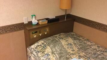A hotel room with a bed, a nightstand with a lamp, a digital clock, and a small table with a tissue box and a spray bottle.