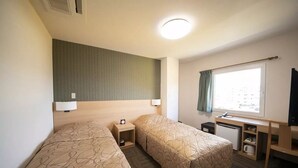Ein Hotelzimmer mit zwei Einzelbetten, einem Schreibtisch, einem Stuhl, einer kleinen Minibar und einem Fenster mit Vorhängen.