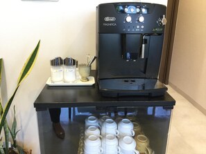Eine De'Longhi MAGNIFICA Kaffeemaschine mit Wasserbehälter, einer Ablage mit Sahne- und Zuckerbehältern und einer Vitrine mit weißen Tassen.