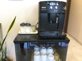 Una cafetera De'Longhi MAGNIFICA con depósito de agua, una bandeja con recipientes para leche y azúcar, y una vitrina con tazas blancas.