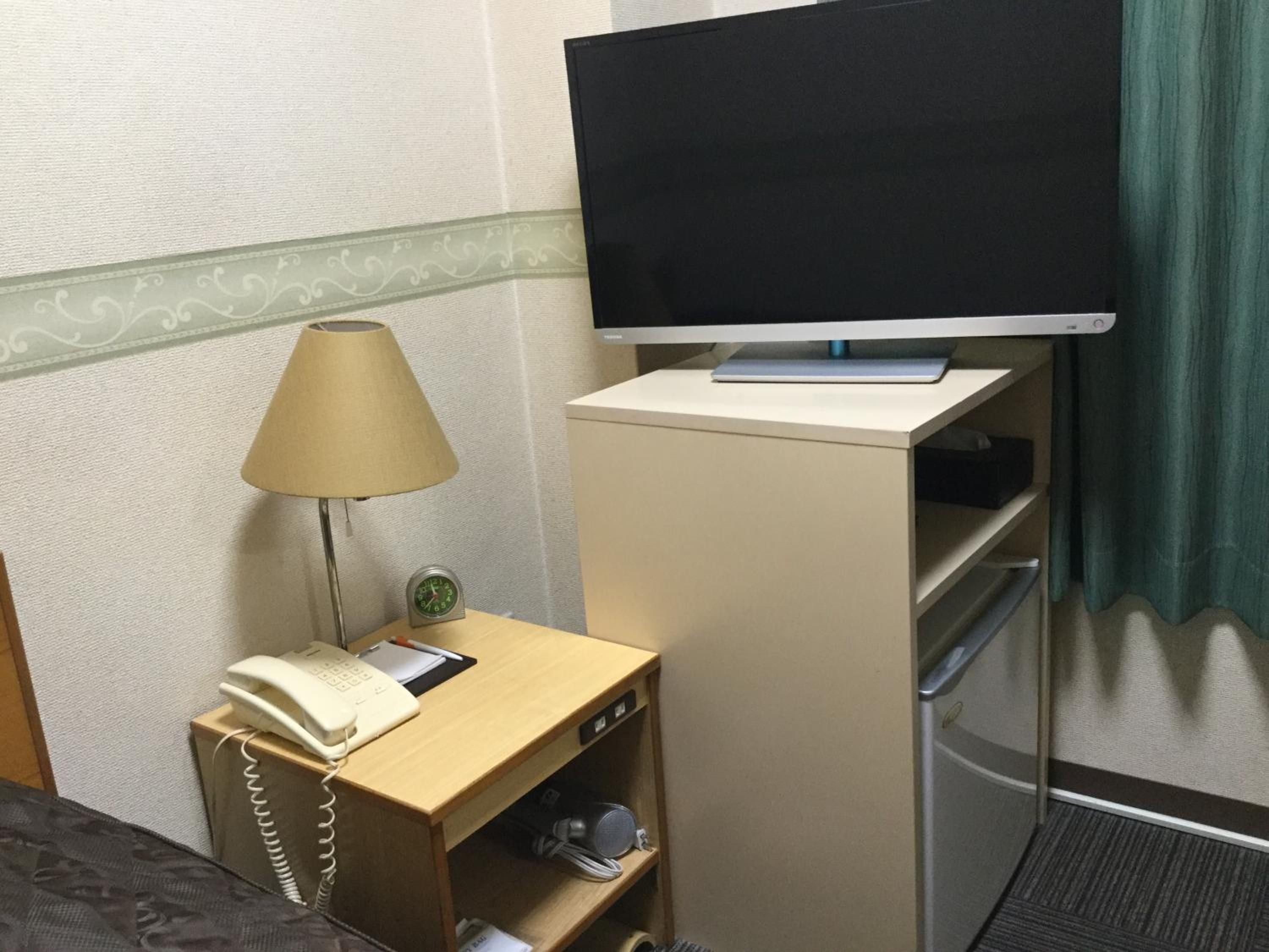 A hotel room with a bed, a desk with a lamp, a television on a stand, and a telephone.