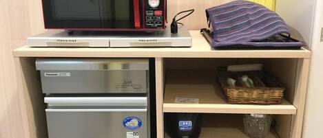 A compact kitchenette with a microwave, refrigerator, and a shelf with a basket and a bag.