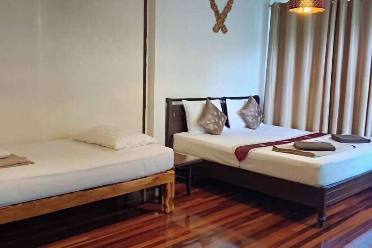 A room with two beds, a wooden floor, a wall-mounted light fixture, and a window with curtains.