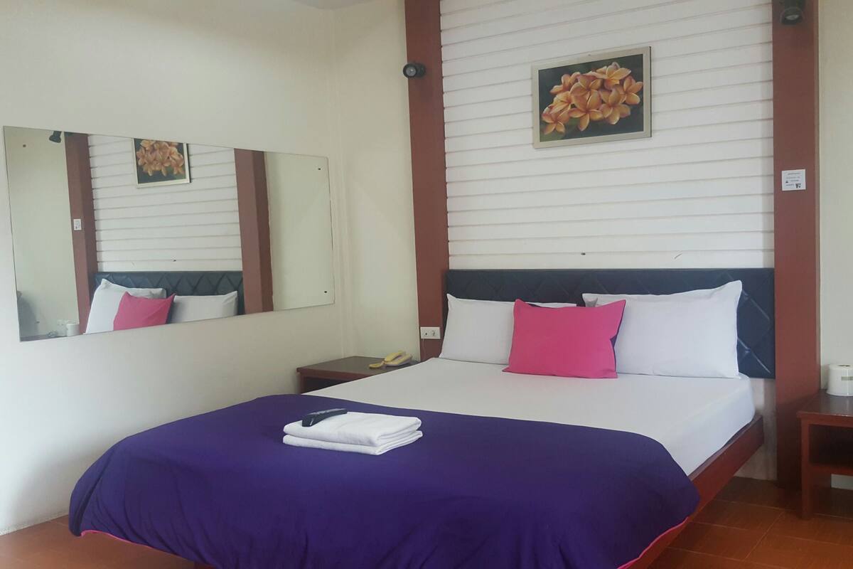 A hotel room with a bed, a bedside table, a nightstand, a mirror, and a wall with a floral painting.
