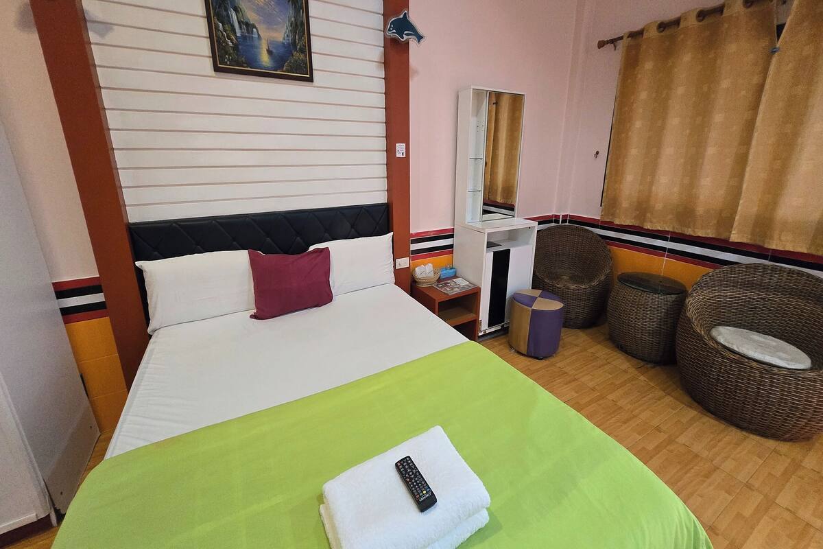 A hotel room with a bed, a green bedspread, a white pillow, a remote control, a mirror, wicker furniture, and a window with curtains.