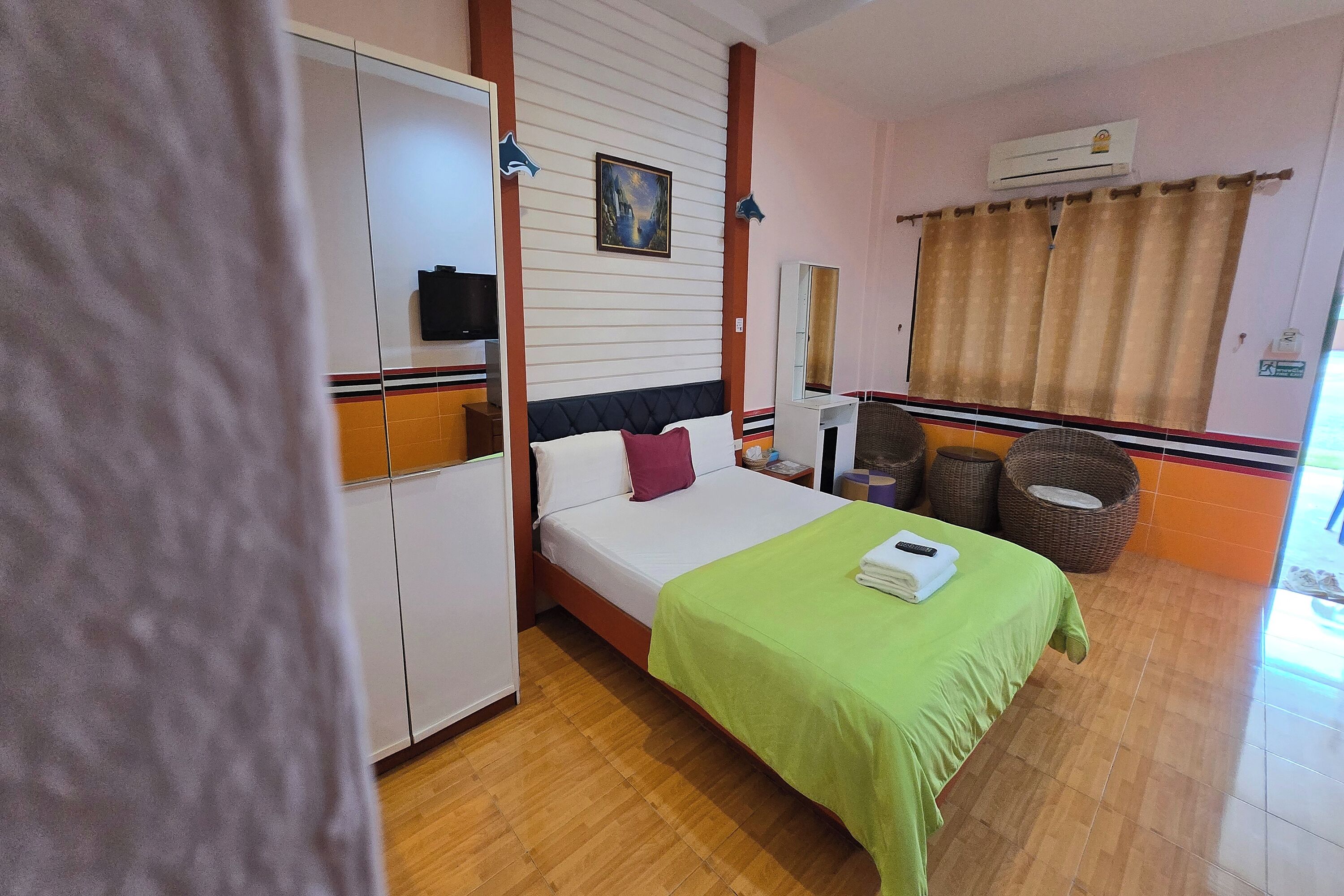 A hotel room with a bed, a wardrobe, a television, a painting, a window with curtains, and a fan.